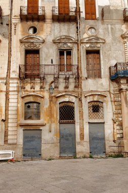 Süslü kemerler, dekoratif demir bir balkon ve ahşap panjurlar içeren yıpranmış bir bina cephesi. Eski duvarlar, desenli alçı ve yıpranmış kapılar klasik cazibe ve kentsel bozulma taşır..