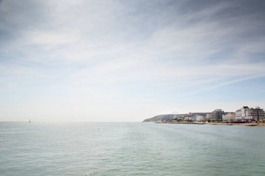 Sakin bir deniz manzarası ufukta bir kıyı kasabası, hafif dalgalar ve uzak bir dönme dolap gösterir. Açık gökyüzü ve köpüklü su sakin, seyahat ve deniz manzarası taşır..