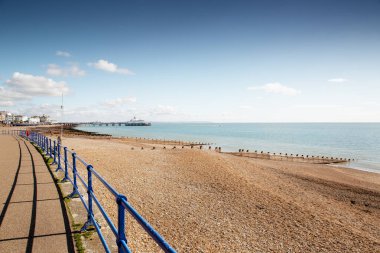 Çakıl taşlı sahil boyunca uzanan mavi korkulukların bulunduğu parlak bir sahil manzarası, sakin bir deniz ve dağınık bulutlu mavi bir gökyüzünün altındaki uzak bir rıhtım..