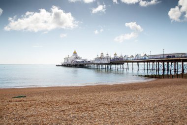 Tarihi Viktorya dönemi iskelesi, çakıllı bir sahilden parlak ve bulutlu bir gökyüzünün altındaki sakin mavi sulara uzanır. Altın kubbeli binalar, sakin dalgaların sahil şeridine ulaşmasıyla cazibe katıyor..