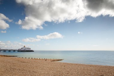 Huzurlu bir sahil manzarası, bir çakıl taşı plajı, ahşap damızlıklar ve dağınık bulutlu, parlak bir gökyüzünün altında sakin mavi sulara uzanan uzak bir rıhtım. Seyahat, boş zaman ve yaz ruh hali görselleri için ideal..