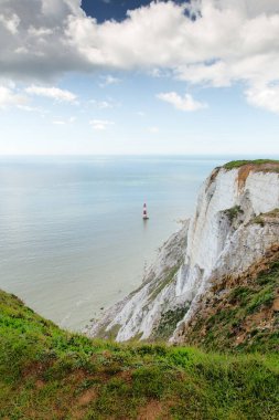 Çimenli uçurumun kenarından dramatik bir manzara beyaz tebeşir uçurumlarına bakar, durgun suda duran tek bir kırmızı-beyaz deniz feneri, parçalı bulutlu mavi gökyüzünün altında. Ada sahili havası ve sakin manzarası.