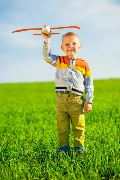 Glücklicher Junge spielt mit Spielzeugflugzeug vor blauem Sommerhimmel und grünem Feldhintergrund. — Stockfoto