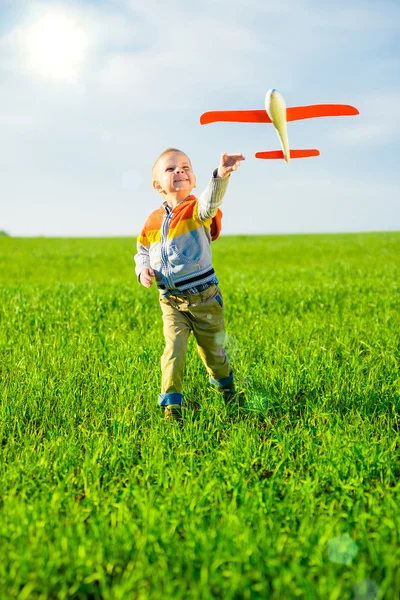 Gelukkige jongen spelen met speelgoed vliegtuig tegen blauwe zomer hemel en groen veld achtergrond. — Stockfoto