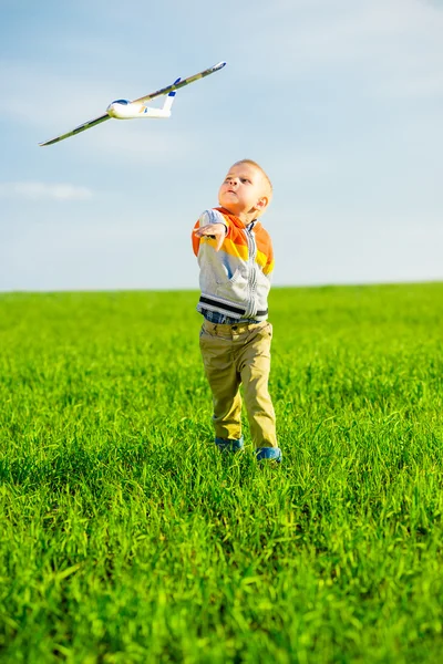 Niño feliz jugando con el avión de juguete contra el cielo azul de verano y el fondo verde del campo . — Foto de Stock