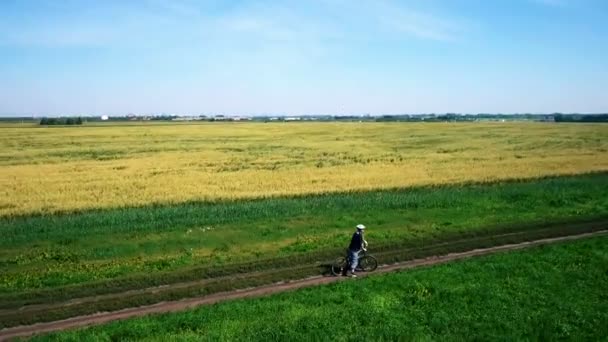 AERIAL: Jovem pedalando de bicicleta na estrada rural através do campo verde e amarelo . — Vídeo de Stock