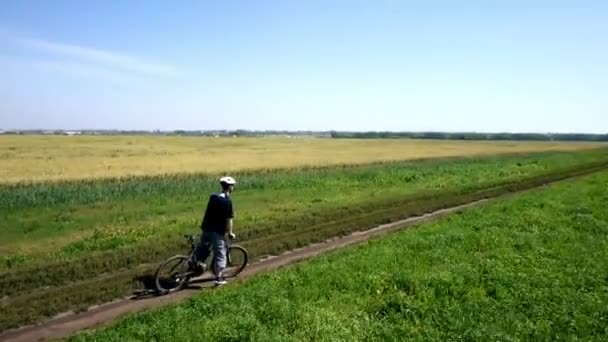 AERIAL: Jovem pedalando de bicicleta na estrada rural através do campo verde e amarelo . — Vídeo de Stock