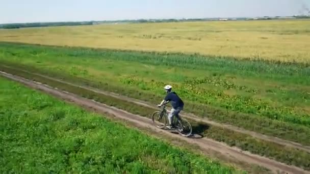 Κεραία: Νεαρός άνδρας ποδηλασία σε ποδήλατο σε αγροτικό δρόμο μέσα από πράσινο και κίτρινο πεδίο. — Αρχείο Βίντεο