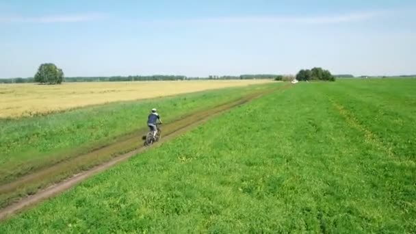 AERIAL: Jovem pedalando de bicicleta na estrada rural através do campo verde e amarelo . — Vídeo de Stock