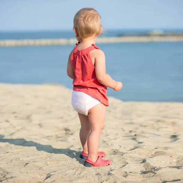 Baby girl in red dress playing on sandy beach near the sea. — Stock Photo, Image