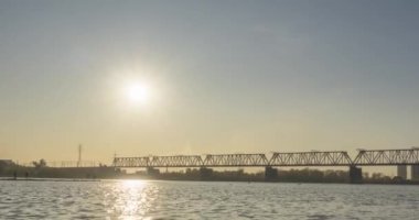 Şehir nehir kıyısının zaman çizelgesi. Güneş ışınları, mavi gökyüzü ve ufukta demiryolu köprüsü. Düz küçük dalgalar, su ve motorlu kaydırak hareketleriyle yaz günbatımı zamanı