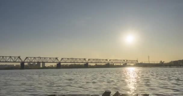 A városi folyópart időrendje. Napsugarak, kék ég és vasúti híd a horizont felett. Nyári naplementekor lapos apróhullámok víz és motoros csúszó mozgás — Stock videók