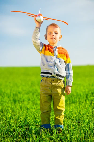Gelukkige jongen spelen met speelgoed vliegtuig tegen blauwe zomer hemel en groen veld achtergrond. — Stockfoto