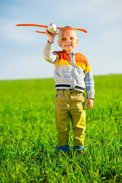 Glücklicher Junge spielt mit Spielzeugflugzeug vor blauem Sommerhimmel und grünem Feldhintergrund. — Stockfoto