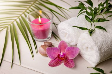 Beyaz ahşap masa üzerinde mum ve pembe orkide çiçekli spa havluları.
