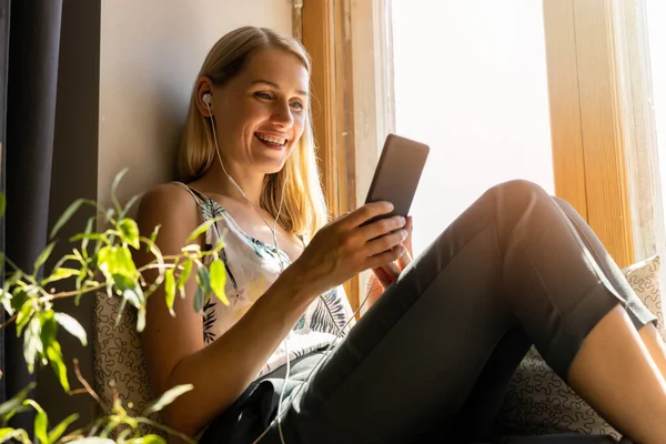 Uśmiechnięta Młoda Kobieta Siedzi Słonecznym Parapecie Używa Smartfona Domu — Zdjęcie stockowe