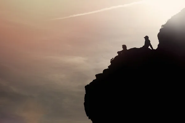 Personas siluetas sentadas en la roca al atardecer — Foto de Stock
