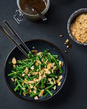 Gri bir masada siyah bir kasede yeşil fasulye. Üst Manzara. Kasede bir kaşık var. Yanında soslu ve bademli kavanozlar var. Kapat.