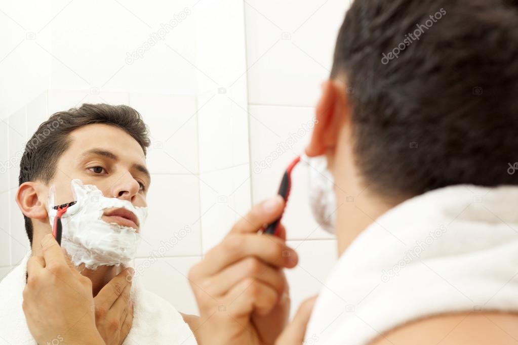 Young man shaving his beard with razor at the bathroom