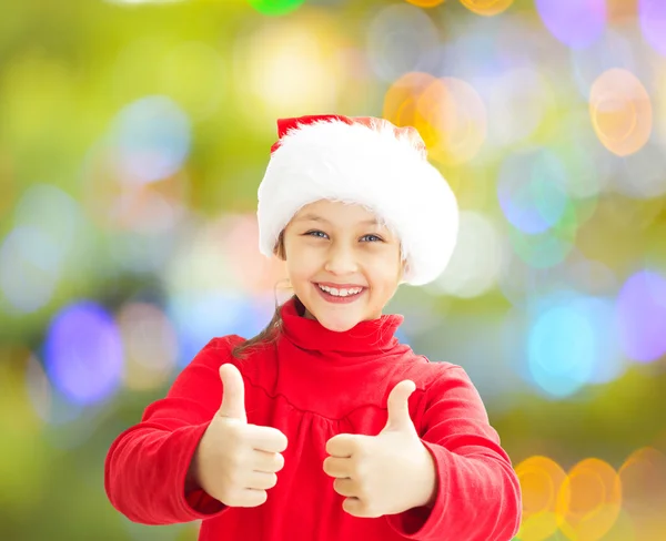 Little girl in a Santa Claus hat shows two thumbs on a colorful — Stock Photo, Image