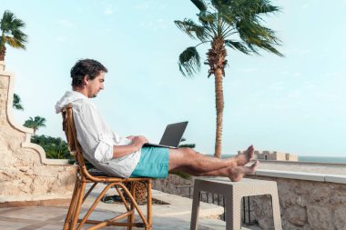 Elinde dizüstü bilgisayarı olan bir adam dinleniyor ve palmiye ağaçlarının arka planında serbest çalışıyor.