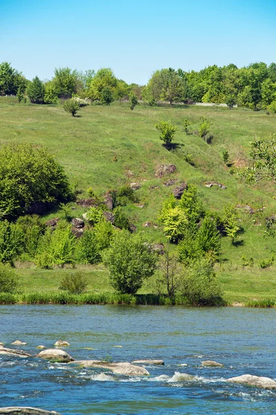 Landscape of grassy hills, trees, river with rock and sky — Stock Photo, Image