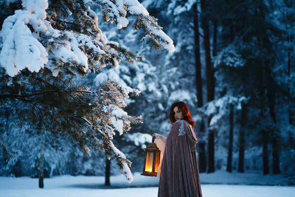 Fantasy cosplay: Young woman with vintage lantern looking at fir tree covered in snow in winter forest. Medieval fairytale concept