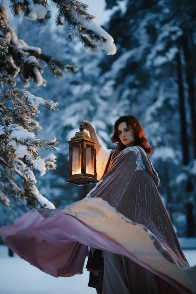Efsanevi fantezi fotoğrafı: genç bir kadın gece vakti karlı ormanda elinde eski model fenerle uçan pelerinini çeviriyor. Ortaçağ peri masalı kostümü konsepti