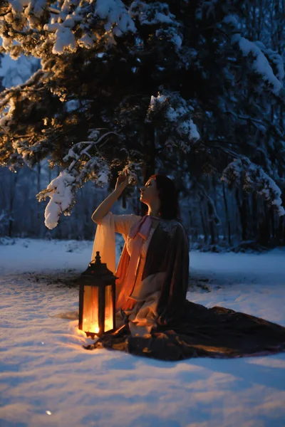 Young woman with vintage lantern sitting under fir tree covered in snow. Medieval fairytale concept