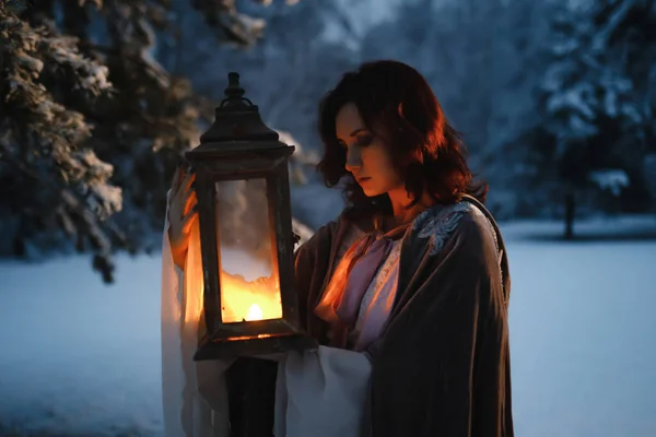 Beautiful fairytale shot of young woman looking at vintage lantern emitting warm light. Medieval fantasy concept Stock Photo