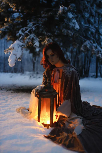 Young woman sitting under fir tree covered in snow looking at vintage lantern. Medieval fairytale concept Royalty Free Stock Images