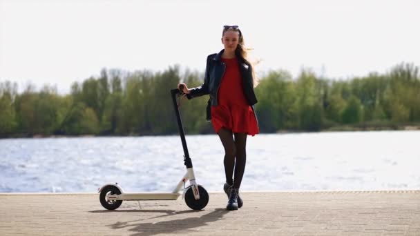 Mladá stylová žena stojí a drží elektrický skútr na promenádě — Stock video