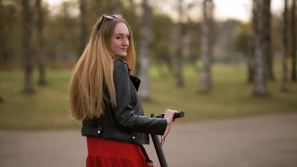 Mladá stylová žena jezdí na elektrickém skútru v městském parku během západu slunce — Stock video
