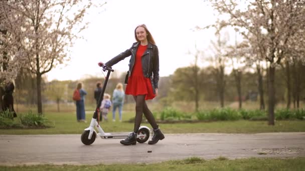 Joven mujer con estilo se para y sostiene scooter eléctrico en el parque de la ciudad — Vídeo de stock