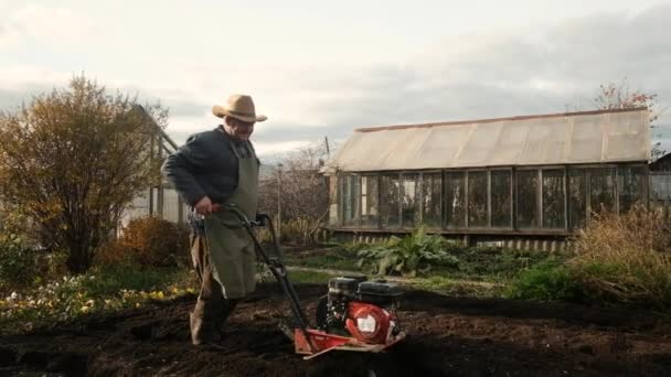 60 세의 농부는 정원에서 모터 재배자 인 트랙터를 사용하여 토양을 경작 한다. 정원가꾸기 개념. 농업 분야의 연금 생활자들을 위한 파트타임으로 일하고 있습니다. 일하고, 건강하고, 활발하고 — 비디오
