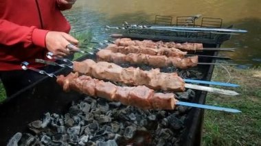 Barbeküyü yakın. Adam ızgarada et kızartıyor. Dışarıda yemek pişirmek. Et, metal şişler üzerinde ızgara dumanına bulanmış. Nehir kıyısındaki doğada dinlenin. kömür