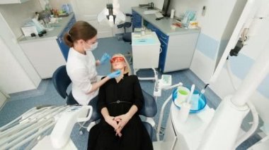 Dişçi, dişçi aletlerini kadın bir müşterinin ağzında tutuyor. Dişçi bir elinde sondayı, diğer elinde de sözlüğü tutuyor. Doktor dişlerin durumunu kontrol ediyor. dolap