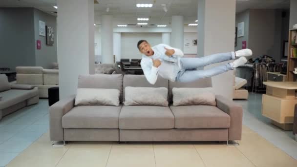 Хлопець стрибає на дивані. Щасливий студент стрибає плащ на дивані в квартирі, будинку, меблевому магазині — стокове відео