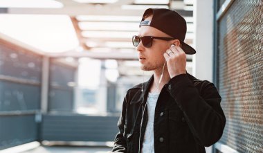 Siyah şapkalı, güneş gözlüklü, şehir günbatımında arka planda akıllı telefon ve kulaklık takan modaya uygun bir adam.