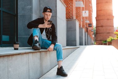 Siyah şapkalı, güneş gözlüklü, şehir günbatımında arka planda akıllı telefon ve kulaklık takan modaya uygun bir adam.