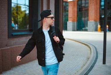 Siyah şapkalı, güneş gözlüklü, şehir günbatımında arka planda akıllı telefon ve kulaklık takan modaya uygun bir adam.