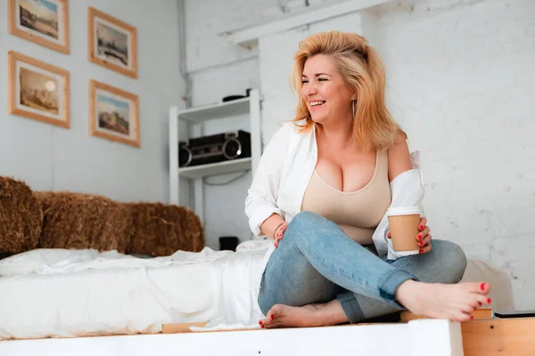 beautiful plus-size girl with big breasts sits on a white floor with coffee. Body positive, diet, curvy figure, sexy, blonde 30 years old, fashion, gluttony, care.