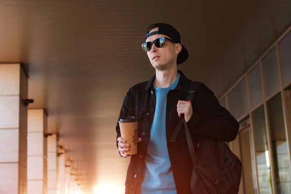 Joven hipster chico de pie calle de la ciudad gorra negra, sosteniendo taza de café. fondo moderno pasillo de cristal de la calle — Foto de Stock