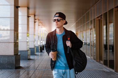 Genç hippi, elinde bir fincan kahve tutan siyah şapkalı, güneş gözlüklü bir şehirde duruyor. Arka planda modern bir sokak cam koridoru var. Sırt çantalı erkek turist ileriye bakıyor.