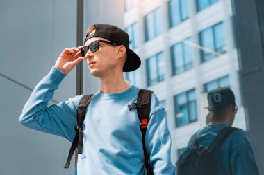 Genç, modaya uygun adam güneş gözlüğü ve sırt çantası öğrenci turist. Arka planda modern cam bina var. Adam iş toplantısına gitmek için acele ediyor. Gelecek vaadeden yaşam tarzı konsepti.