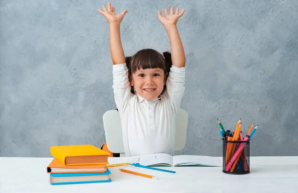 De vuelta a la escuela. Niña linda colegiala sentada en un escritorio en la habitación. El chico está aprendiendo y haciendo su tarea. La chica es emocionalmente feliz con las manos en alto — Foto de Stock