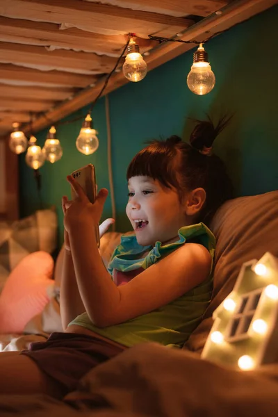 Niña feliz con un peinado divertido sentado en la habitación de los niños en su litera sosteniendo el teléfono inteligente — Foto de Stock