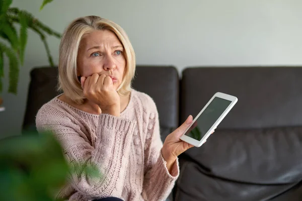 Mulher Madura triste sentado no sofá e usando comprimido digital. Navegando internet sobre tablet em casa. — Fotografia de Stock