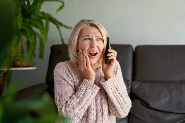 Triste mujer mayor en una llamada telefónica sentada en un sofá en la sala de estar — Foto de Stock