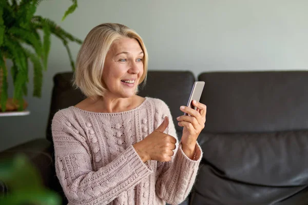 Mulher sênior feliz usando telefonema sentado em um sofá na sala de estar em uma casa — Fotografia de Stock